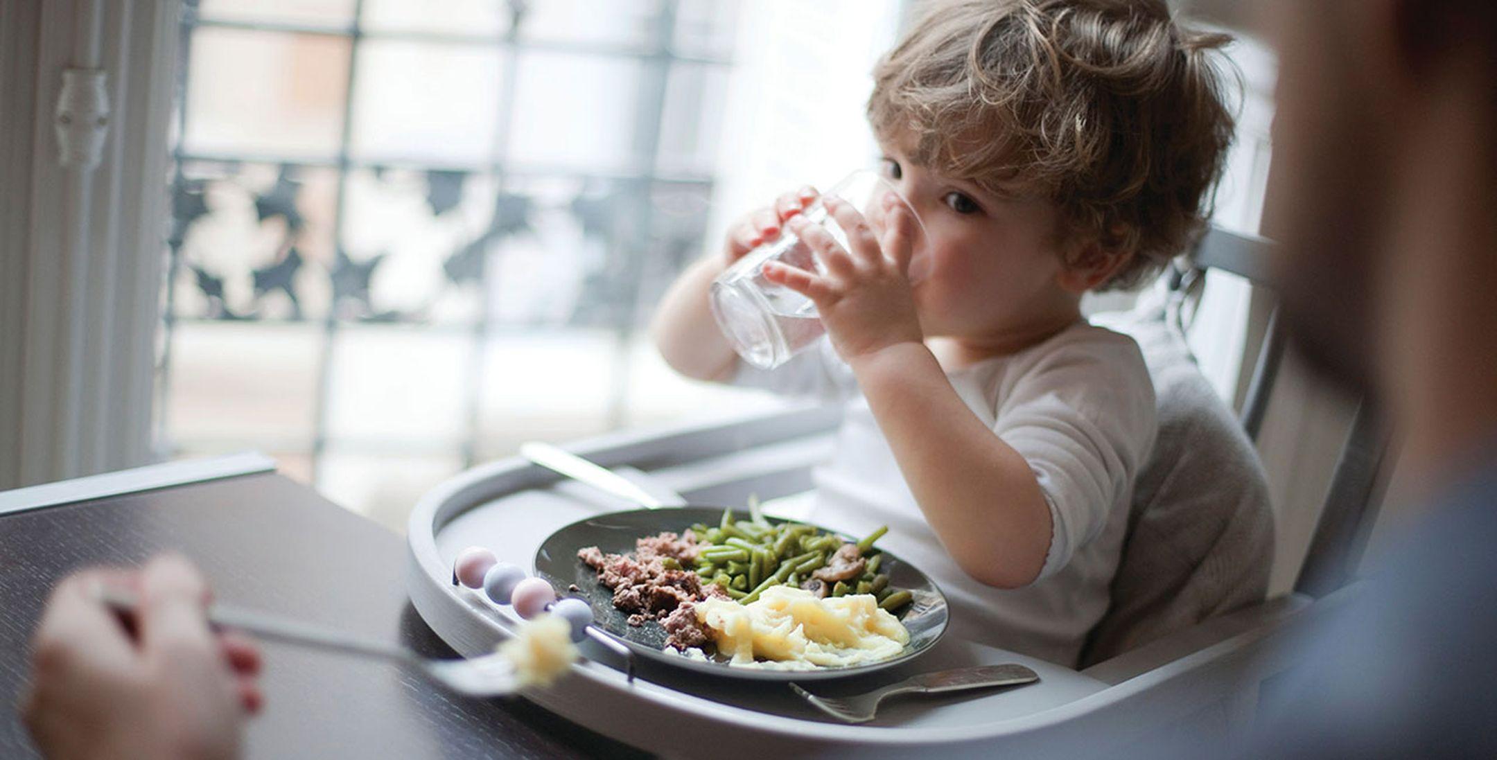 Small child sitting in a high chair with a plate full of food while drinking from a glass that he's holding with two hands.